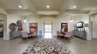 This single-floor community space makes a strong first impression with its open, airy lobby and lounge area. The interior is anchored by a distinctive coffered ceiling with recessed lighting and dual ceiling fans, lending a refined architectural character to the room. Large French doors at the far end of the space flood the interior with natural light while providing a seamless visual connection to the outdoors.

The lounge area is thoughtfully arranged with multiple seating clusters, each featuring red and white checkered upholstered chairs paired with dark bistro-style tables — creating a warm and inviting atmosphere for gathering. A bold, multicolored circular-patterned area rug serves as the centerpiece of the space, adding a lively and modern contrast to the neutral cream and beige wall tones and tile flooring.

Framed artwork on both sides of the room contributes to the welcoming ambiance, while decorative accents such as a large floral vase and a sideboard with a lamp add warmth and personality. The symmetrical layout and generous proportions of the space make it well-suited for socializing and community activities. Overall, the space strikes a comfortable balance between casual and polished, offering a functional yet visually engaging environment.