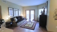 This single-story living space showcases a well-appointed living room that combines comfort and classic style. The room features warm hardwood flooring that extends throughout, providing a cohesive and inviting foundation. A pair of French doors serves as a focal point, allowing natural light to filter in through slatted shutters while offering access to the outdoors. Flanking a dark, plush sofa are two framed wall prints that add a decorative touch to the soft sage-green walls. The living area is anchored by a patterned area rug with a bold border, layered beneath an ornate wrought-iron and glass coffee table. A dark wood console or cabinet positioned to the right adds both storage and visual contrast to the lighter tones of the room. The space is thoughtfully arranged to maximize flow and functionality, with decorative accents including a classic clock and potted greenery contributing to the overall warmth. A side window further enhances the brightness of the room. The overall ambiance is relaxed yet polished, reflecting a traditional aesthetic with carefully curated furnishings and a comfortable, livable design.