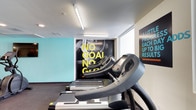 This fitness center offers a well-appointed workout environment designed to motivate and support an active lifestyle. The space features a clean, contemporary aesthetic with bright white ceilings accented by linear fluorescent lighting that ensures the area is well-lit throughout. Bold motivational graphics and large-scale wall murals serve as focal points, featuring empowering phrases that set an energetic tone for the facility. A vibrant teal accent wall adds a pop of color, contributing to the dynamic and modern atmosphere. The cardio section is equipped with a row of 7000 Series treadmills, providing members with quality equipment for their exercise routines. Natural light filters in through a window at the rear of the space, adding to the bright and open feel of the room. The flooring is finished in a dark, durable material well-suited for a high-traffic fitness environment. Overall, the fitness center strikes a balance between functionality and motivating design, making it an inviting space for individuals looking to pursue their health and wellness goals.