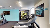 This fitness center offers a well-appointed workout environment designed to motivate and support an active lifestyle. The space features a clean, contemporary aesthetic with bright white ceilings accented by linear fluorescent lighting that ensures the area is well-lit throughout. Bold motivational graphics and large-scale wall murals serve as focal points, featuring empowering phrases that set an energetic tone for the facility. A vibrant teal accent wall adds a pop of color, contributing to the dynamic and modern atmosphere. The cardio section is equipped with a row of 7000 Series treadmills, providing members with quality equipment for their exercise routines. Natural light filters in through a window at the rear of the space, adding to the bright and open feel of the room. The flooring is finished in a dark, durable material well-suited for a high-traffic fitness environment. Overall, the fitness center strikes a balance between functionality and motivating design, making it an inviting space for individuals looking to pursue their health and wellness goals.