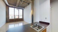 This single-floor loft-style space showcases a compelling blend of industrial character and modern functionality. The exposed concrete ceiling with visible ductwork and utility pipes gives the space a raw, urban aesthetic, while original wooden support beams add warmth and structural authenticity. Track lighting runs along the ceiling, providing flexible, directed illumination throughout.

The open main area features polished hardwood floors that reflect natural light pouring in through large, multi-pane factory-style windows. These oversized windows frame views of the surrounding brick streetscape, enhancing the industrial ambiance while keeping the interior bright and airy. The high ceilings amplify the sense of openness and volume throughout the space.

A functional kitchenette area includes a stainless steel sink set into a dark countertop, offering practical utility within the open-plan layout. A long corridor connects the various sections of the space, lined with white walls and accented by a classic six-panel glass-pane interior door that allows light to pass through while maintaining defined zones. The polished floors continue seamlessly throughout the hallway, tying the entire space together with a cohesive, clean finish. Overall, this space offers a distinctive industrial-loft character well-suited for creative or professional use.