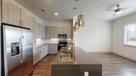 This single-floor residence showcases a contemporary design with clean lines and a neutral palette throughout. The open-concept kitchen serves as the centerpiece, featuring flat-front cabinetry in a warm gray-taupe finish, a white subway tile backsplash, and a full suite of stainless steel appliances including a French door refrigerator with ice maker, electric range, and microwave. A generous kitchen island with a dark quartz countertop and undermount stainless steel sink anchors the space, illuminated by a trio of smoked glass pendant lights that add a refined industrial accent. Wood-look plank flooring flows seamlessly through the kitchen and living areas, providing a warm, cohesive foundation.

The bedroom is bright and airy, benefiting from multiple windows with exterior views and access to a private door, all fitted with blinds. A ceiling fan adds functional comfort. An adjacent walk-in closet offers multi-tiered shelving and hanging rods set over plush carpeting for dedicated storage. The bathroom presents a clean, upscale aesthetic with a granite-topped vanity, wood-toned cabinetry, wood-look tile flooring, and a full soaking tub surrounded by floor-to-ceiling dark wood-grain tile surround.