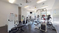 This fitness center is a comprehensively equipped single-level facility designed to support a full range of workout routines. The space features white walls, dark rubber flooring, and a bright, airy atmosphere created by large windows that line one side of the room, offering views of the surrounding outdoor environment and flooding the interior with natural light. Ceiling fans and recessed fluorescent lighting ensure consistent illumination and air circulation throughout.

The cardio zone is well-stocked with treadmills, elliptical trainers, stationary bikes, and a stair climber, several of which are positioned directly in front of the large windows—an ideal setup for an engaging workout experience. Wall-mounted televisions are positioned throughout for entertainment during exercise sessions.

The strength training area features Magnum-branded cable and resistance machines, a multi-function cable tower, and adjustable benches to support a variety of exercises. A dedicated free weights alcove includes a tiered dumbbell rack with a full range of weights, a workout bench, and large wall mirrors that visually expand the space and allow for form monitoring.

Open shelving provides organized storage for accessories and informational materials. The combination of natural light, ample equipment variety, and thoughtful layout makes this fitness center a functional and inviting space for daily exercise.