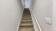This efficiently designed one-bedroom, one-bathroom unit offers a practical layout across a single floor, accessible via a carpeted staircase with white-painted railings that conveys a clean, simple aesthetic. The neutral gray and beige color palette used throughout creates a cohesive, understated ambiance.

The bedroom is a comfortable, carpeted space with soft beige tones, a six-panel white door, and access to the hallway landing with white baluster railings. A dedicated closet provides additional storage. A standout feature of the main living area is a set of French doors that open to an outdoor balcony, allowing natural light to fill the space and offering outdoor access.

The bathroom includes a full-size soaking tub with a surrounding surround enclosure in a clean white finish. The dedicated laundry room is equipped with washer hookups, a water heater, and wood-look flooring, providing functional utility space. White cabinetry visible in the 3D scan suggests a kitchen with ample storage. Overall, the unit offers a well-organized, functional layout suited for efficient everyday living.