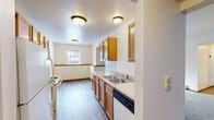 Located at 1921 Parkcrest Dr SW in Wyoming, Michigan, this single-floor unit offers four bedrooms and two bathrooms within a well-organized layout. The galley-style kitchen is a functional highlight, featuring warm honey-toned wood cabinetry, light countertops, a double-basin stainless steel sink, a dishwasher, and a white electric range — all set atop durable wood-look flooring that continues into the adjoining dining or living area. Natural light filters in through a window at the far end, enhancing the open feel of this combined space. The two bathrooms maintain a consistent, clean aesthetic: the full bath includes a shower-tub combination, wood-toned vanity, and medicine cabinet mirror, while the second bath features a single vanity with mirror and a grab bar for added accessibility. Each of the four bedrooms is finished with neutral carpet, warm wood baseboard trim, and individual windows that bring in natural light. One bedroom includes a baseboard heater, ensuring comfort throughout the seasons. A dedicated laundry closet houses a stacked washer and dryer unit alongside built-in shelving, adding significant in-unit convenience. Hallways connect all spaces efficiently, completing this straightforward and functional single-level layout.