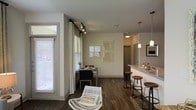 This single-floor residence offers a well-designed open-concept layout that seamlessly connects the living, dining, and kitchen areas into one cohesive living space. Dark hardwood flooring runs throughout, providing a warm contrast against the neutral gray-toned walls and white trim that define the interior palette.

The kitchen features a granite-topped breakfast bar with bar stool seating, pendant lighting overhead, and a clean, modern aesthetic. It opens directly into the dining area, which is illuminated by natural light streaming through large windows dressed with white curtains on black rod hardware. Track lighting provides additional ambiance and flexibility in the main living area.

The dining space comfortably accommodates a table with seating, while a woven accent chair adds a casual, relaxed touch to the living zone. Decorative wall art in neutral tones adds visual interest without overwhelming the space. The layout promotes an easy flow between functional areas, making it ideal for both everyday living and entertaining.

Overall, the space presents a modern, understated aesthetic with thoughtful design details, ample natural light, and a cohesive material palette that creates a comfortable and inviting atmosphere throughout.