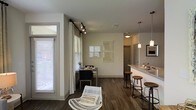This single-floor residence offers a well-designed open-concept layout that seamlessly connects the living, dining, and kitchen areas into one cohesive living space. Dark hardwood flooring runs throughout, providing a warm contrast against the neutral gray-toned walls and white trim that define the interior palette.

The kitchen features a granite-topped breakfast bar with bar stool seating, pendant lighting overhead, and a clean, modern aesthetic. It opens directly into the dining area, which is illuminated by natural light streaming through large windows dressed with white curtains on black rod hardware. Track lighting provides additional ambiance and flexibility in the main living area.

The dining space comfortably accommodates a table with seating, while a woven accent chair adds a casual, relaxed touch to the living zone. Decorative wall art in neutral tones adds visual interest without overwhelming the space. The layout promotes an easy flow between functional areas, making it ideal for both everyday living and entertaining.

Overall, the space presents a modern, understated aesthetic with thoughtful design details, ample natural light, and a cohesive material palette that creates a comfortable and inviting atmosphere throughout.