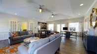 This single-floor space offers a thoughtfully designed open-concept layout that seamlessly blends comfort and functionality. The main lounge area is anchored by plush gray sofas arranged around a rustic wooden coffee table, creating a welcoming gathering spot. Overhead, industrial-style ceiling fans with pendant lighting illuminate the space while contributing to its relaxed, contemporary aesthetic. Rich, dark hardwood flooring runs throughout, adding warmth and visual continuity across all areas.

A kitchen area is visible toward the back, equipped with stainless steel appliances, including a refrigerator, and features a striking wood-accent wall that adds a rustic, textural element to the space. Adjacent to the kitchen, a casual seating nook with leather chairs provides an intimate spot for smaller conversations or relaxed reading.

Natural light pours in through multiple large windows dressed with patterned curtains, keeping the interior bright and airy. A dedicated co-working zone along one wall features a long wooden desk with desktop computers, making it a practical workspace for residents or guests. The yellow accent wall near the entrance and the white-paned glass cabinetry add character and charm. Overall, the space balances a homey, inviting atmosphere with versatile, multi-use functionality.