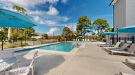 Located at 1910 E First St in Gulf Shores, Alabama, this outdoor amenity area is centered around a generously sized swimming pool set within a well-maintained, single-level complex. The pool itself features clean, clear blue water with convenient step entry points and is complemented by an attached wading area, making it suitable for a range of swimmers. The expansive concrete sun deck surrounding the pool is furnished with rows of white lounge chairs and teal market umbrellas, providing ample space for relaxation in the coastal Alabama sun. A dedicated shade structure on one side of the deck offers a covered retreat from the heat. The area is enclosed by black iron fencing, ensuring both safety and a clean aesthetic. Mature pine, palm, and mixed subtropical trees line the perimeter, framing the space with natural greenery and providing additional shade. An accessory storage building is positioned at the edge of the deck, adding functional utility to the space. The surrounding residential buildings are visible from the pool area, reflecting the community-oriented nature of this amenity. The overall atmosphere is bright, open, and inviting, well-suited to the warm Gulf Coast climate and outdoor lifestyle that defines the Gulf Shores area.