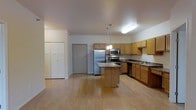 This single-floor space is designed with an open-concept layout that seamlessly integrates the kitchen and living areas, creating a spacious and functional environment. The kitchen is a central highlight, featuring a full suite of stainless steel appliances, including a refrigerator, electric range, and built-in microwave. Light wood cabinetry lines the walls, offering generous storage, and is complemented by granite-style countertops that add a polished aesthetic. A center island with matching countertop and pendant lighting above serves as both a practical workspace and a visual focal point within the room. The flooring throughout is light hardwood, contributing to the bright and airy feel of the space. A double-door closet near the entry provides additional storage. The open floor plan between the kitchen and adjacent living area allows for flexible furniture arrangement and easy flow between spaces. Natural light, combined with overhead fluorescent lighting, keeps the space well-illuminated. The neutral color palette of white walls and warm wood tones creates a clean, welcoming atmosphere throughout.