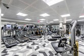 This fitness center offers a spacious, single-level layout designed to accommodate a comprehensive range of workout activities. The space is immediately distinguished by its bold black, white, and grey geometric-patterned carpet tiles, which create a visually dynamic foundation throughout the entire floor area.

The cardio zone is prominently featured along one side of the facility and is lined with a generous row of modern treadmills, elliptical trainers, stair climbers, and stationary bikes, each equipped with individual digital display screens. Full-length mirrors run along the walls in this section, enhancing the sense of openness and allowing users to monitor their form during exercise.

The strength and functional training area occupies the opposite end of the space and includes cable machines, a multi-station cable tower, free weights, weight plates, and benches. A structural column anchors the center of this zone without impeding the flow of movement throughout the room.

The facility is accessed through glass-paneled entry doors framed by stainless steel accents, with steps leading into the main floor. Overhead, a recessed drop ceiling fitted with evenly spaced flush panel lights provides consistent, bright illumination across the entire space. The overall ambiance is clean, modern, and functional.