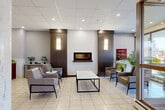 Located at 1032 King St W in Kingston, Ontario, this residential building lobby offers a well-appointed and functional entry experience. The generously sized single-floor space is finished with light ceramic tile flooring throughout, complemented by a drop ceiling with recessed pot lighting that keeps the area bright and inviting.

A comfortable lounge area anchors one side of the lobby, furnished with contemporary leather armchairs, side tables, and a central coffee table arranged around a sleek wall-mounted linear fireplace framed by dark wood accent panels with warm sconce lighting — creating a welcoming atmosphere for residents and guests. Tall glazed entry doors and a large window frontage allow ample natural light to fill the space and provide views to the surrounding streetscape.

Prominent cylindrical concrete columns add a distinctive architectural character to the open-plan layout. Toward the rear, two stainless steel elevators provide vertical access throughout the building, alongside a secondary access door. A dedicated mailroom area along one wall features a full bank of individual resident mailboxes and a parcel locker station. A carpeted hallway corridor extends further into the building, connecting the lobby to additional amenities and residential units. The overall ambiance is clean, modern, and well-maintained.