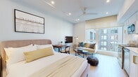 This well-appointed studio is a masterclass in efficient, elegant design. The entry hallway sets a refined tone, featuring light hardwood flooring, recessed lighting, a decorative gold-framed mirror, and curated wall art with matching gold frames — offering an inviting introduction to the space.

The open main area seamlessly blends the sleeping, living, and kitchen zones. The sleeping area is anchored by a generously sized upholstered bed, complemented by a wall-mounted flat-screen TV and a compact wooden desk with a bentwood chair — ideal for work or leisure. A ceiling fan with recessed lighting overhead adds both function and style.

The living area features a neutral loveseat sofa, a round glass coffee table, and a floor lamp, all positioned to take full advantage of the expansive floor-to-ceiling windows that flood the space with natural light and offer views of the surrounding cityscape.

The kitchen is outfitted with white shaker cabinetry, a striking marble-look backsplash, white quartz countertops, brushed gold hardware and faucet, a stainless steel undermount sink, and a full suite of stainless steel appliances.

The bathroom is finished with warm travertine-style tile, a floating white vanity with gold fixtures, and clean, modern lines throughout. A mirrored walk-in closet provides ample storage, completing this compact yet highly functional space.