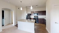 This single-story residence at 6310 S Watson Rd in Grand Prairie, Texas offers a well-appointed layout designed for comfortable everyday living. The kitchen serves as a central highlight, featuring rich dark-stained cabinetry paired with light granite countertops that extend to a generously sized white island with a built-in double sink. Overhead, pendant lights and a track lighting system illuminate the space, which is further enhanced by notably tall ceilings that lend an open, airy quality. Black appliances, including a refrigerator, electric range, and built-in microwave, complement the cabinetry for a cohesive aesthetic. Hard-surface flooring runs throughout the kitchen area, transitioning to plush carpet in the living and sleeping areas. The bedroom is well-sized, receiving natural light through dual windows fitted with blinds, and is equipped with a ceiling fan. A dedicated walk-in closet provides substantial storage, with wood shelving built in for organization. An arched hallway connects the bedroom to the rest of the unit. The bathroom includes a full tub-shower combination with white tile surround, a dark-cabinetry vanity with ample under-sink storage, and a large mirror above the sink, completing the functional and cohesive design of the space.