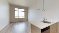 This single-floor unit showcases a clean, contemporary design with light wood-tone flooring running throughout all areas, creating a cohesive and airy aesthetic. The open-concept layout connects the kitchen and living area seamlessly, maximizing the sense of space. The kitchen is outfitted with stainless steel appliances, including a refrigerator, range, and microwave, complemented by white upper cabinetry and light wood lower cabinets. A peninsula with a white quartz-style countertop and undermount sink extends into the living area, with two industrial-style pendant lights hanging above. Track lighting overhead further illuminates the kitchen and hallway. The living area is generously proportioned, with crisp white walls and a large picture window that frames views of surrounding treetops, filling the space with natural light. The bedroom is equally well-lit, featuring its own large window with the same outdoor views, offering a bright and calm retreat. The bathroom is finished with consistent wood-tone flooring, a light wood vanity with a white countertop and integrated sink, a large mirror, and a full bathtub with a shower, maintaining the unit's clean and modern character throughout.