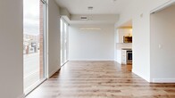 This efficiently designed 1-bedroom, 1-bathroom unit offers a clean, modern aesthetic throughout its single-floor layout. The open-concept living area is immediately impressive, anchored by expansive floor-to-ceiling windows that draw in abundant natural light and offer views of the surrounding streetscape. A contemporary wave-form pendant light fixture adds a stylish focal point above the living space, complementing the warm hardwood-style flooring that runs consistently throughout the main areas.

The kitchen is positioned adjacent to the living area in a galley-style configuration, featuring crisp white cabinetry, stainless steel appliances — including a built-in microwave, range, and dishwasher — and light countertops that reinforce the unit's bright, cohesive palette.

The bedroom is a well-proportioned, private retreat with mirrored sliding closet doors that enhance the sense of space, along with its own access to natural light. The bathroom area is connected to a utility space that houses an in-unit stacked washer and dryer — a practical and convenient feature.

Throughout the unit, white walls, light tile flooring in the utility corridor, and consistent trim details create a clean, modern environment. The overall design emphasizes functionality and brightness, making the most of the available footprint with a well-organized and contemporary layout.