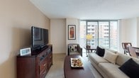 Located on the seventh floor at 7620 Old Georgetown Road in Bethesda, Maryland, this apartment offers a thoughtfully designed single-level layout with warm, neutral tones throughout. The living room serves as the heart of the home, featuring floor-to-ceiling windows that flood the space with natural light and open to a private balcony with urban views of the surrounding Bethesda skyline and greenery. The adjacent dining area is defined by an elegant chandelier and is well-suited for entertaining. The kitchen is efficiently laid out with warm wood cabinetry, granite-style countertops, tile flooring, and white appliances. The primary bedroom is generously sized with plush carpeting, a window with sheer curtains, and ample closet space. It connects to a spacious en-suite bathroom featuring a long vanity with dual mirrors, warm wood cabinetry, and a full tub-shower combination. The second bedroom is equally comfortable, offering carpeted flooring, a large window, and a cozy seating area. A second full bathroom with a tub-shower combo and clean white finishes serves this bedroom. Throughout, the apartment maintains a cohesive, warm aesthetic with beige and taupe tones, creating a calm and inviting atmosphere.