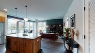 This thoughtfully designed single-floor urban residence blends contemporary aesthetics with warm, mid-century modern touches throughout. The open-concept layout seamlessly connects the kitchen, dining, and living areas, creating a generous and inviting communal space flooded with natural light from expansive floor-to-ceiling windows that frame striking city views.

The kitchen is a standout feature, outfitted with rich walnut-toned cabinetry, a white subway tile backsplash, stone countertops, stainless steel appliances, and a large island with seating — ideal for both everyday cooking and entertaining. Globe pendant lights add a warm, curated ambiance overhead.

The living room is anchored by a bold deep-green accent wall hosting a full built-in shelving unit and media center, complemented by an architectural round column and large windows that pour in natural light and urban scenery.

The primary bedroom offers a serene retreat with a dramatic green accent wall, a floor-to-ceiling window with city views, and access to a well-appointed en-suite bathroom featuring dual vanities, a glass-enclosed walk-in shower with white tile, and a spacious walk-in closet. A second bedroom is equally bright, with large windows overlooking the cityscape. A second full bathroom with a soaking tub and tiled surround completes the layout. Wide-plank hardwood flooring flows throughout, tying the entire residence together cohesively.