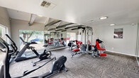 Located in Houston, TX, this fitness center at 1620 Bay Area Boulevard offers a well-equipped exercise space designed to support a comprehensive workout routine. The room features a striking vaulted ceiling with exposed dark wood beams against a white shiplap surface, lending the space an architectural character that sets it apart from a typical gym environment. Large windows along multiple walls flood the interior with natural light and provide views of the surrounding outdoor pool area and landscaping, creating an open and energizing atmosphere. The flooring consists of durable, patterned carpet tiles throughout. Floor-to-ceiling mirrors are strategically positioned along the walls, visually expanding the space and allowing users to monitor their form during exercise. The fitness center is outfitted with a variety of cardio machines, including stationary bikes, a treadmill, and an elliptical, alongside a multi-station strength training machine with red upholstered pads and benches, and additional resistance equipment. A wall-mounted television is also present for entertainment during workouts. Recessed and flush-mount lighting supplements the ample natural light, ensuring the space remains bright and functional at all hours. The overall ambiance is clean, modern, and purpose-built for fitness.