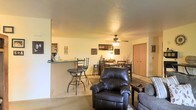 This single-floor residence is designed with an open-concept layout that seamlessly connects the main living areas, creating a cohesive and functional living environment. The living room serves as the central gathering space, furnished with a leather recliner and a plush sofa, anchored by a ceiling fan with a light fixture that provides both comfort and illumination. Adjacent to the living room, a raised breakfast bar with bar stool seating offers a casual dining option, separated from the kitchen by a half-wall partition that maintains an open feel while defining each space. A dedicated dining area sits just beyond, accommodating a full dining table and chairs, making it well-suited for everyday meals and entertaining. The interiors feature neutral-toned walls and light carpeting throughout, contributing to a warm and inviting ambiance. Wall art and decorative accents add personality to the space. A closeted door is visible in the background, suggesting additional storage or room access. The layout flows efficiently across the single floor, making the space practical and easy to navigate.