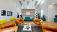 Located at 961 Marcus Dr in Newport News, Virginia, this single-floor community clubhouse is a thoughtfully designed amenity space that blends bold color and contemporary style. The central gathering area commands attention with soaring ceilings, recessed lighting, and large windows that invite abundant natural light. A striking statement wall featuring a vivid, multicolored abstract pattern serves as the focal point, complemented by a teal sofa, rich cognac leather armchairs, and a sleek coffee table arranged atop a textured area rug — creating a welcoming and visually dynamic lounge. An adjacent seating nook offers a quieter retreat, furnished with charcoal upholstered chairs, small accent tables, and curated wall art for a more intimate atmosphere. The kitchenette is a standout feature, boasting deep teal cabinetry, bold hexagonal tile backsplash, open wood shelving, pendant lighting, and a breakfast bar with colorful seating — fully equipped for residents to enjoy refreshments. A dedicated business center, accessible through a glass-paned door, provides a functional workspace. Throughout the space, light wood-look flooring, two-tone wainscoting, and carefully chosen décor elements tie each area together into a cohesive, modern aesthetic that is both practical and inviting.