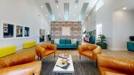 Located at 961 Marcus Dr in Newport News, Virginia, this single-floor community clubhouse is a thoughtfully designed amenity space that blends bold color and contemporary style. The central gathering area commands attention with soaring ceilings, recessed lighting, and large windows that invite abundant natural light. A striking statement wall featuring a vivid, multicolored abstract pattern serves as the focal point, complemented by a teal sofa, rich cognac leather armchairs, and a sleek coffee table arranged atop a textured area rug — creating a welcoming and visually dynamic lounge. An adjacent seating nook offers a quieter retreat, furnished with charcoal upholstered chairs, small accent tables, and curated wall art for a more intimate atmosphere. The kitchenette is a standout feature, boasting deep teal cabinetry, bold hexagonal tile backsplash, open wood shelving, pendant lighting, and a breakfast bar with colorful seating — fully equipped for residents to enjoy refreshments. A dedicated business center, accessible through a glass-paned door, provides a functional workspace. Throughout the space, light wood-look flooring, two-tone wainscoting, and carefully chosen décor elements tie each area together into a cohesive, modern aesthetic that is both practical and inviting.
