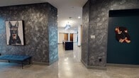 Located on W Corporation St in Durham, NC, this single-floor amenity space showcases a thoughtfully designed interior that blends industrial and contemporary aesthetics. Polished concrete floors run throughout, reflecting light from recessed ceiling fixtures and creating a cohesive, modern foundation across all areas.

The main lobby and common areas feature dramatic textured stone-clad columns with a brushed, dark finish, acting as bold architectural anchors. A spacious lounge area boasts soaring ceilings, with pendant lighting suspended above a central seating arrangement. Built-in shelving and curated artwork add character, while a vertical wood slat partition elegantly defines separate zones without fully closing them off.

The club room offers comfortable lounge seating and floor-to-ceiling windows with sliding glass doors opening to an outdoor balcony, providing panoramic views of the surrounding Durham cityscape. A kitchenette is appointed with quartz countertops, sage-green cabinetry, a black undermount sink, penny-tile backsplash, and vibrant citrus-themed wallcovering.

A dedicated meeting room features a round white table, deep teal accent walls, textured wood paneling, and curated wall art. Private workstations tucked into teal-painted alcoves offer focused work areas. Barn-style sliding doors and elevator access complete this well-appointed communal space.