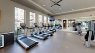 This expansive fitness center is thoughtfully designed to accommodate a full range of workout routines in a bright, welcoming environment. The space is organized into distinct zones, each tailored to a specific type of training. The main workout area is generously sized, featuring rows of commercial-grade treadmills and cardio equipment positioned along a wall of large windows that flood the space with natural light and offer views of the surrounding outdoor areas. Full-length mirrors line one wall, enhancing the sense of openness while allowing users to monitor their form. A dedicated strength training zone houses a comprehensive selection of weight machines and cable equipment, complemented by additional mirrors and mounted flat-screen televisions throughout. A separate cycling studio is set apart with a bold accent wall, stationary bikes, and a mounted TV, creating an energetic atmosphere ideal for focused training sessions. The studio is also equipped with a beverage cooler and towel station, adding a convenient, amenity-forward touch. Recessed lighting, ceiling fans, tray ceilings, and durable flooring throughout give the facility a polished, professional feel. Resistance bands, stability balls, and functional training accessories round out the offerings, making this a comprehensive fitness destination.