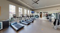 This expansive fitness center is thoughtfully designed to accommodate a full range of workout routines in a bright, welcoming environment. The space is organized into distinct zones, each tailored to a specific type of training. The main workout area is generously sized, featuring rows of commercial-grade treadmills and cardio equipment positioned along a wall of large windows that flood the space with natural light and offer views of the surrounding outdoor areas. Full-length mirrors line one wall, enhancing the sense of openness while allowing users to monitor their form. A dedicated strength training zone houses a comprehensive selection of weight machines and cable equipment, complemented by additional mirrors and mounted flat-screen televisions throughout. A separate cycling studio is set apart with a bold accent wall, stationary bikes, and a mounted TV, creating an energetic atmosphere ideal for focused training sessions. The studio is also equipped with a beverage cooler and towel station, adding a convenient, amenity-forward touch. Recessed lighting, ceiling fans, tray ceilings, and durable flooring throughout give the facility a polished, professional feel. Resistance bands, stability balls, and functional training accessories round out the offerings, making this a comprehensive fitness destination.