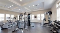 This single-floor fitness center offers a comprehensive workout environment designed to accommodate a wide range of exercise routines. The space is defined by its elegant tray ceilings with recessed lighting and ceiling fans, which contribute to a bright, comfortable atmosphere throughout the workout area. Large windows fitted with blinds line the walls, allowing abundant natural light to fill the room while providing views of the surrounding exterior. The durable rubber flooring provides a safe, cushioned surface ideal for both cardio and strength training activities. The cardio section is well-stocked, featuring multiple treadmills, elliptical machines, and a stair climber, offering users a variety of aerobic workout options. A multi-station cable machine serves as the centerpiece for strength training, complemented by a free weights rack with dumbbells, a weight bench, and a barbell rack with plates. Wall-mounted flat-screen televisions are positioned for easy viewing during workouts. A glass-paneled entry door with transom window provides direct outdoor access, enhancing the sense of openness. The space is thoughtfully laid out to ensure ample room between equipment, supporting a comfortable and functional fitness experience.