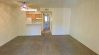 This single-floor residence offers a practical and straightforward layout designed for comfortable everyday living. The main living area is finished with wall-to-wall carpeting, creating a warm and cohesive atmosphere throughout. A ceiling fan with an integrated light fixture is positioned overhead, providing both ventilation and ambient lighting to the space.

The kitchen is situated adjacent to the living area and is defined by a peninsula-style breakfast bar that serves as a natural divider between the two spaces. It is equipped with warm-toned wood cabinetry, a white refrigerator, and a white range, offering a clean and functional cooking environment. A ventilation duct is visible in the ceiling, ensuring proper airflow throughout the unit. The kitchen also benefits from a light fixture that keeps the workspace well-illuminated.

A hallway extending from the main living area leads toward additional rooms at the rear of the unit, with natural light visible at the far end, suggesting a window or exterior-facing door. The walls throughout are finished in a neutral off-white tone, complementing the textured ceiling and contributing to a bright, open feel. The overall layout is efficiently organized, making good use of the available space across the single floor.