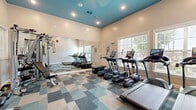 Located at 961 Seven Oaks Blvd in Smyrna, Tennessee, this fitness center offers a well-equipped workout space designed to support a range of exercise routines. The room features soaring ceilings painted in a bold teal-blue accent color, creating a vibrant and energizing atmosphere complemented by recessed lighting and natural light streaming through multiple large windows with views of the surrounding greenery.

The flooring consists of stylish geometric-patterned carpet tiles in shades of teal, gray, and beige, adding both visual interest and comfort underfoot. A full-length wall mirror runs along one side of the room, effectively amplifying the sense of space and allowing for proper form checks during workouts.

The cardio section is well-appointed with multiple True-brand treadmills and elliptical machines, each equipped with individual display screens. Strength training is supported by a tiered dumbbell rack stocked with a comprehensive range of free weights, a weight bench, and a multi-function cable and resistance machine. The dark-paneled entry door adds a clean architectural contrast to the light-toned walls.

Overall, the fitness center strikes a balance between functional utility and modern design, providing residents with a dedicated, comfortable space for daily exercise.