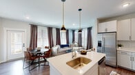 This thoughtfully designed 3-bedroom, 2-bathroom single-story residence offers a cohesive open-concept floor plan that maximizes every square foot. The kitchen is a standout feature, outfitted with white shaker cabinetry, stainless steel appliances including a refrigerator, microwave, and range, a subway tile backsplash, and a quartz-topped island with bar seating. Dual pendant lights with brass accents add a modern touch overhead. The kitchen flows seamlessly into the dining area, which benefits from a glass-paneled exterior door that invites natural light, and onward into the living room, where multiple windows dressed in layered drapes and wood blinds create a bright, welcoming atmosphere. Warm-toned hardwood-style flooring runs throughout the main living areas, lending visual continuity. A clean, white hallway connects the bedrooms and bathrooms to the main living spaces. The primary bedroom is generously proportioned, featuring large windows with ample natural light and a ceiling fan. The bathrooms are finished with white vanities, quartz countertops, large mirrors, and wood-style flooring. A dedicated laundry room houses a full-size top-load washer and dryer. The overall aesthetic is modern and neutral, with clean lines and contemporary finishes throughout.