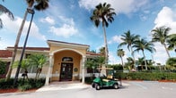 This single-story building showcases classic Florida Mediterranean architecture, characterized by a warm yellow stucco exterior, a red tile roofline, and elegant arched entryway supported by white columns. The grand double-door entrance is framed by decorative archwork, lending a refined and welcoming appearance to the facade. A covered portico provides a shaded approach to the front doors, ideal for Florida's sunny climate. The exterior is beautifully complemented by lush, mature tropical landscaping, including towering palm trees and manicured hedges that create a verdant, resort-like atmosphere. Paved walkways and a spacious parking area in front of the building offer convenient access. The overall design and setting evoke a relaxed yet polished Florida lifestyle, consistent with a community clubhouse, recreation center, or similar gathering facility. Natural light and open skies enhance the bright, airy ambiance of the surroundings, while the tropical foliage provides both shade and visual appeal. The combination of Mediterranean architectural details and lush greenery gives this space a distinctive, welcoming character well-suited to a warm, coastal environment.