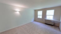 This single-floor space presents a clean, minimalist layout characterized by neutral wall tones and wall-to-wall carpeting in a soft beige hue, contributing to a warm and understated aesthetic. The main living area is notably open, offering a generous sense of space with smooth, white ceilings and crisp white baseboards that frame the room cohesively.

A wall-mounted sconce provides ambient accent lighting along one side of the room, complementing the natural light that enters through two windows positioned along the far wall. These windows feature roll-down shades and allow views of surrounding greenery, lending a pleasant connection to the outdoors. The windows also contribute to a well-lit interior environment throughout the day.

A wall-mounted air conditioning unit is positioned beneath one of the windows, offering a practical climate control solution. The overall design of the space is straightforward and functional, with a neutral color palette throughout that provides a versatile backdrop suitable for a variety of furnishing styles. The room's open layout and soft finishes create a calm, livable atmosphere.
