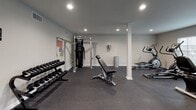 This fitness space is thoughtfully designed to support a full range of exercise activities within a single-floor layout. The room features neutral gray walls, recessed lighting, and durable rubber-speckled flooring throughout, creating a practical and focused environment for physical training.

The free weights area includes a multi-tiered dumbbell rack stocked with a complete set of hex dumbbells, accompanied by an adjustable weight bench ideal for a variety of strength exercises. A vertical medicine ball storage rack holds an assortment of weighted balls, adding functional training options to the space. A cable machine provides additional resistance training versatility, and a reference chart mounted on the wall offers exercise guidance.

The cardio section is well-appointed with two TRUE treadmills, two elliptical trainers, and a stationary bike, all positioned near windows fitted with blinds that allow natural light to filter in. Wall-mounted flat-screen televisions serve both the cardio and strength areas, enhancing the workout experience. Structural support columns are integrated cleanly into the layout, and two access doors contribute to smooth circulation throughout the space.