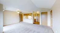 This single-floor space features an open-concept layout that seamlessly connects the living area to the kitchen. The living room is finished with neutral wall-to-wall carpeting that adds warmth underfoot, and a ceiling fan with integrated lighting provides both comfort and illumination. A vaulted ceiling section adds an airy sense of volume to the space.

The kitchen is accessible via a pass-through counter, offering a practical and social connection between the cooking and living areas. It is outfitted with warm honey-toned wood cabinetry, a white refrigerator, and a sink positioned at the pass-through. The kitchen transitions into a hardwood-floored area beyond, where additional cabinetry is visible, suggesting a dedicated dining or utility space nearby.

Warm wood-toned doors are consistent throughout the unit, contributing to a cohesive aesthetic. Two doors are visible from the main living area — one on the far left and one on the right — likely leading to private rooms or storage. The overall palette of soft whites, warm neutrals, and natural wood tones creates a clean, functional, and inviting atmosphere throughout the space.