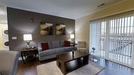 Located at 1 Park Lane Boulevard in Cherry Hill, New Jersey, this single-floor residence offers a thoughtfully designed living space with a warm and contemporary aesthetic. The living area serves as the heart of the home, featuring rich hardwood flooring, crown molding, and a neutral color palette accented by a bold charcoal feature wall. Large sliding glass doors fitted with vertical blinds allow generous natural light to fill the room while providing access to outdoor views. The space comfortably accommodates a full seating arrangement, complemented by tasteful wall art and ambient table lighting that contribute to an inviting atmosphere. The combination of warm and cool tones throughout creates a balanced, modern ambiance suitable for both relaxing and entertaining. Crown molding along the ceiling adds a refined architectural detail that elevates the overall design. The layout is practical and well-proportioned, offering a seamless flow within the single-level floor plan. Overall, this Cherry Hill residence blends classic detailing with contemporary interior design, making it a comfortable and visually appealing space in a well-established New Jersey community.