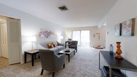 This single-floor residence centers around a spacious living area designed with comfort and functionality in mind. The living room is finished with wall-to-wall plush carpeting in a warm neutral tone, contributing to the overall soft and welcoming ambiance of the space. White walls and a clean white ceiling amplify the natural light that streams in through a large sliding glass door, which opens to what appears to be a balcony or outdoor area, providing both ventilation and an extended sense of openness. The sliding door is fitted with vertical blinds, offering adjustable privacy and light control. A centrally located ceiling vent ensures efficient climate management throughout the room. The layout comfortably accommodates a full seating arrangement, including a sofa and accent chairs positioned around a central coffee table, making it well-suited for both relaxing and entertaining. Wall art adds a subtle decorative touch against the neutral backdrop. The overall design is clean and understated, with an emphasis on open space, natural light, and a neutral color palette that offers a versatile foundation for a variety of interior styles.