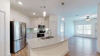 This well-appointed single-floor residence offers a thoughtfully designed open-concept layout that seamlessly connects the kitchen and living areas. The kitchen is a standout feature, showcasing a large curved island with granite countertops and an undermount sink, white shaker-style cabinetry, a soft aqua glass tile backsplash, and a full suite of stainless steel appliances including a French door refrigerator, electric range, and microwave. Pendant lighting above the island adds a polished finishing touch. A dedicated pantry provides convenient additional storage. The living area extends naturally from the kitchen, benefiting from abundant natural light through large windows and featuring warm hardwood-style flooring and a ceiling fan. The bedroom is generously sized, carpeted for comfort, and includes a ceiling fan and a walk-in closet with built-in shelving. The en-suite bathroom is finished with dual undermount sinks set into granite countertops, modern brushed nickel fixtures, and a large mirror, creating a clean and functional space. Throughout the residence, crisp white walls, recessed lighting, and consistent high-quality finishes contribute to a bright, contemporary ambiance.