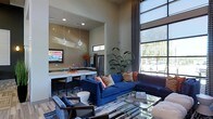 This single-floor space showcases a contemporary open-concept design that effortlessly blends living, dining, and kitchen areas into one cohesive environment. The living area is anchored by a bold navy blue sectional sofa accented with vibrant patterned throw pillows, complemented by sleek glass coffee tables and stylish accent chairs upholstered in a geometric print. A large area rug in cool blue tones defines the seating zone and adds warmth to the light wood-style flooring beneath.

One of the most striking architectural features is the dramatic floor-to-ceiling window wall that floods the interior with abundant natural light and offers expansive outdoor views. Soaring ceilings amplify the sense of openness and grandeur throughout the space. Patterned drapery panels frame the oversized windows, adding texture and visual depth.

The kitchen is seamlessly integrated into the open floor plan, featuring a white countertop island with bar seating, pendant lighting, and stainless steel appliances including a full-size refrigerator. A wall-mounted flat-screen television is positioned for easy viewing from both the kitchen and living areas. Lush tropical potted plants are thoughtfully placed to bring natural elements indoors, enhancing the bright and airy ambiance of this well-appointed space.