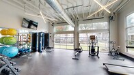 Located at 4500 Sherwood Common Blvd in Baton Rouge, Louisiana, this well-appointed fitness center offers a thoughtfully designed workout environment across a single floor. The exercise room is the heart of the space, distinguished by soaring ceilings with exposed white structural beams and industrial-style ductwork that lend it a modern, open-air aesthetic. Linear pendant lighting runs across the ceiling, complementing the abundant natural light that pours in through large floor-to-ceiling glass garage-style doors along one wall, as well as additional windows that frame views of the surrounding greenery and outdoor grounds.

The exercise room is equipped with a comprehensive selection of fitness equipment, including cardio machines such as ellipticals, a rowing machine, and a treadmill, as well as strength-training stations, a cable machine, a leg press, a rack of dumbbells spanning a wide weight range, and a colorful collection of medicine and stability balls. A wall-mounted flat-screen television adds a multimedia element for an enhanced workout experience. The durable speckled rubber flooring throughout provides both safety and functionality.

A dedicated bathroom off the main exercise area features ADA-compliant accessibility, including grab bars, a tile floor in slate tones, and clean white shiplap-style wainscoting, ensuring convenience and inclusivity for all users.