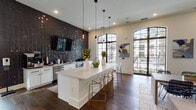 This single-floor amenity space showcases a sophisticated blend of modern and Mediterranean-inspired design elements. The main gathering area commands attention with dramatic vaulted ceilings supported by dark-stained exposed wood beams, complemented by striking geometric pendant chandeliers that draw the eye upward. Large arched windows and full-height glass doors flood the interior with natural light while offering views of an outdoor pool and lush landscaping beyond.

Rich hardwood flooring flows throughout the space, anchoring an open-plan layout that seamlessly connects the lounge, dining, and kitchenette areas. The focal point of the kitchen zone is a sleek white island with a quartz countertop, surrounded by modern acrylic and brass bar seating. A dramatic floor-to-ceiling textured dark tile accent wall houses a mounted flat-screen TV and a coffee and beverage station, adding bold contrast to the crisp white cabinetry.

The lounge area features curated contemporary artwork, arched wall niches with decorative sconces, and thoughtfully arranged seating that creates intimate conversation zones. A large patterned area rug anchors the dining table, which is styled with woven black leather chairs beneath a sculptural chandelier. The entry corridor introduces the space with art-adorned walls and natural light streaming through glass-paneled doors, setting an inviting, gallery-like tone throughout.