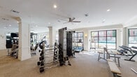 This fitness center in Houston, TX, is a thoughtfully designed, single-floor facility that combines functionality with refined aesthetics. The space features crisp white walls adorned with classic crown molding and decorative pilasters, creating an upscale ambiance. Neutral-toned carpeted flooring runs throughout, complementing the clean, modern interior.

Large, black-framed multi-pane windows line the perimeter, flooding the space with abundant natural light and offering views of an outdoor pool area and surrounding greenery. Wall sconces provide warm accent lighting, enhancing the inviting atmosphere.

The cardio zone is well-appointed with treadmills, stationary cycling bikes, and a StairMaster stair climber, all positioned to take advantage of the outdoor views. A dedicated strength training area includes a multi-station cable machine, a variety of resistance machines, and a fully stocked dumbbell rack displayed in front of a floor-to-ceiling mirror. Additional benches and functional training accessories round out the equipment offerings.

A ceiling fan centrally positioned overhead aids in air circulation, while posted workout charts on the wall provide guidance for users. The open, spacious layout ensures ample room to move freely between stations, making this fitness center a comprehensive and comfortable environment for a full range of exercise routines.