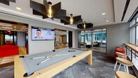 Located in the heart of downtown Raleigh at 511 Fayetteville St, this contemporary amenity floor delivers a dynamic blend of social, recreational, and relaxation spaces. The entry corridor makes an immediate impression with a bold geometric-patterned floor, a textured ceiling treatment, and vibrant orange sectional seating anchored by a large-format display wall.

The game room features a full-size billiards table beneath striking angular acoustic ceiling panels and pendant lighting, with views into adjacent lounge areas. A dedicated lounge area showcases a circular tray ceiling with integrated lighting, floor-to-ceiling windows that flood the space with natural light, and comfortable curved seating arrangements ideal for gatherings or casual meetings.

The kitchen and dining area is outfitted with a substantial dark-topped island with bar seating, pendant lighting, built-in appliances, wood cabinetry, and a large mounted display. Multiple dining configurations accommodate both intimate and group settings.

Colorful lounge nooks offer bold yellow armchairs, built-in window seating, and direct access to the outdoor terrace. The terrace itself features vibrant orange outdoor sofas, glass railings, and sweeping panoramic views of the Raleigh skyline, providing a striking urban backdrop. Throughout, the space balances energetic design elements with functional, well-appointed amenities.