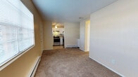 This single-floor space offers a practical and comfortable layout that connects the main living area to a well-equipped kitchen. The living room features wall-to-wall carpet in a neutral tan tone, providing a warm and understated foundation for the space. Natural light filters in through a window fitted with blinds, brightening the room alongside baseboard heating units positioned along the walls, ensuring year-round comfort. The textured popcorn ceiling adds a classic residential character throughout.

The kitchen is visible from the living area and is outfitted with stainless steel appliances, including a gas range and refrigerator. A patterned tile backsplash introduces a subtle decorative element, while under-cabinet lighting and a ceiling fan with a light fixture add both function and ambiance. The kitchen floor transitions to a hard surface, distinguishing it from the carpeted living space.

Two doors are visible — one appearing to lead to a closet and another to an additional room or hallway — suggesting a thoughtful, efficient floor plan. Baseboard heating units are present in multiple areas, contributing to a consistently comfortable interior environment throughout the unit.