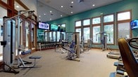 This fitness center is thoughtfully designed to support a comprehensive workout experience across multiple dedicated zones. The main exercise floor features distinctive teal-painted walls accented by warm wood-trimmed windows that fill the space with natural light and offer views of the surrounding outdoors. Recessed lighting and ceiling fans complement the bright, energizing atmosphere throughout.

The strength training area is generously outfitted with a full rack of dumbbells, multiple cable and selectorized machines, and adjustable benches, providing options for a wide range of resistance exercises. Full-length mirrors line the walls, enhancing the sense of openness and allowing for proper form checks during workouts. Multiple wall-mounted flat-screen televisions are positioned throughout for entertainment.

The cardio zone features a row of treadmills, elliptical trainers, and stair climbers from True Fitness, all positioned to take advantage of the large windows and outdoor views. Adjacent to the main floor, a separate studio room with warm hardwood flooring houses spinning bikes, stability balls, step platforms, a heavy bag, and an interactive fitness on-demand screen — ideal for group classes or individual training sessions. The vaulted ceiling in the entry area adds architectural grandeur, creating an inviting and motivating environment for all fitness levels.