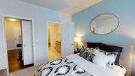 This single-floor residence offers a clean, modern aesthetic with a warm and inviting atmosphere. The bedroom serves as a standout feature, showcasing soft blue accent walls that contrast beautifully against crisp white trim and neutral light gray walls. Plush wall-to-wall carpeting adds comfort underfoot, while a warm bedside lamp casts a welcoming glow throughout the room. A decorative round mirror with a gold frame adorns the accent wall, adding a contemporary touch to the space. The room comfortably accommodates a large bed with stylish, patterned bedding, complemented by a wooden nightstand. Direct access to the en-suite bathroom is provided through a classic white paneled door. The bathroom features wood-toned cabinetry, a vanity with sink, and a toilet, all finished with light-toned flooring that maintains a cohesive, airy feel. A separate door in the bedroom leads to what appears to be a spacious closet, offering generous storage. The overall design of the space balances practicality with style, making it a comfortable and functional living environment.