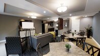This well-appointed single-floor space offers an open-concept layout that seamlessly blends a comfortable lounge, dining areas, and a fully equipped kitchen into one cohesive environment. The lounge area is anchored by a stylish patterned sofa accented with warm throws, complemented by sleek floor lamps that provide a soft, inviting ambiance. Multiple dining tables with dark crossback chairs are arranged throughout the space, accommodating both intimate gatherings and larger group settings. The kitchen features dark cabinetry, stainless steel appliances including a full-size refrigerator and microwave, and is partially separated from the main area by a handsome wooden column detail that adds architectural character. Recessed lighting is thoughtfully distributed across the tray ceiling, while elegant drum pendant light fixtures serve as focal points overhead, casting a warm, welcoming glow throughout. The neutral grey wall tones and contrasting dark furnishings create a sophisticated, contemporary aesthetic. Granite-topped surfaces add a refined touch, while decorative mirrors and greenery contribute to the polished, carefully curated atmosphere of the space.