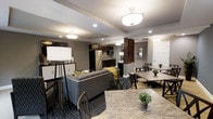 This well-appointed single-floor space offers an open-concept layout that seamlessly blends a comfortable lounge, dining areas, and a fully equipped kitchen into one cohesive environment. The lounge area is anchored by a stylish patterned sofa accented with warm throws, complemented by sleek floor lamps that provide a soft, inviting ambiance. Multiple dining tables with dark crossback chairs are arranged throughout the space, accommodating both intimate gatherings and larger group settings. The kitchen features dark cabinetry, stainless steel appliances including a full-size refrigerator and microwave, and is partially separated from the main area by a handsome wooden column detail that adds architectural character. Recessed lighting is thoughtfully distributed across the tray ceiling, while elegant drum pendant light fixtures serve as focal points overhead, casting a warm, welcoming glow throughout. The neutral grey wall tones and contrasting dark furnishings create a sophisticated, contemporary aesthetic. Granite-topped surfaces add a refined touch, while decorative mirrors and greenery contribute to the polished, carefully curated atmosphere of the space.